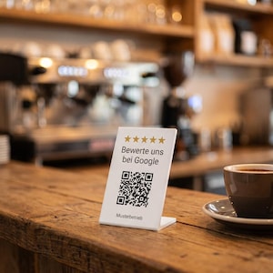 Könnte beinhalten: Ein weißes, L-förmiges Acrylschild mit fünf goldenen Sternen, dem Text "Bewerte uns bei Google", einem schwarz-weißen QR-Code und dem Text "Musterbetrieb" am unteren Rand, platziert auf einer Holzoberfläche neben einer Kaffeetasse.