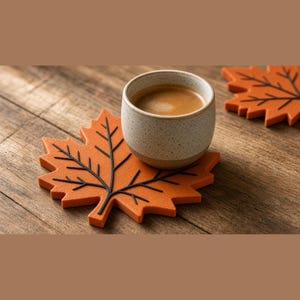 Pode incluir: Uma caneca de cerâmica bege salpicada cheia de café castanho assenta num porta-copos laranja em forma de folha de ácer com detalhes de veios pretos. Um segundo porta-copos de folha laranja idêntico é parcialmente visível no canto superior direito.