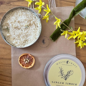 May include: A brown paper bag with a handle, two metal tins, a dried orange slice, and yellow flowers. One tin contains a light-colored granular substance. The other tin has a yellow label with the text "GINGER TIMBER".