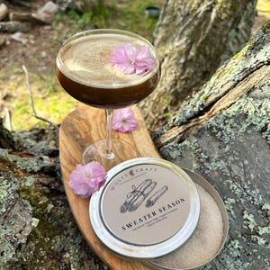 May include: A cocktail in a stemmed glass with a pink flower garnish, resting on a wooden board. A tin with the text "SWEATER SEASON" and cinnamon illustrations sits nearby. The cocktail is dark brown with a foamy top.