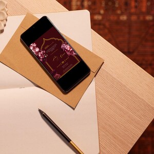 May include: A smartphone displays a burgundy wedding invitation with floral accents, resting on a brown envelope. A notebook, pen, and decorative elements are arranged on a wooden surface, creating an elegant scene.