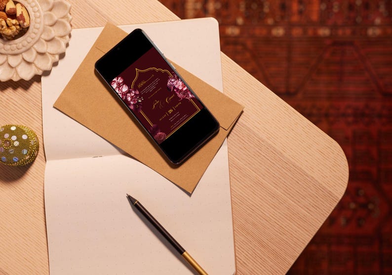 May include: A smartphone displays a burgundy wedding invitation with floral accents, resting on a brown envelope. A notebook, pen, and decorative elements are arranged on a wooden surface, creating an elegant scene.