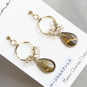 以下が含まれることがあります： クリアビーズのアクセントと涙型のペンダントが付いたゴールドトーンのイヤリング。ペンダントは緑と金のデザインです。イヤリングは、日本語のテキストが入った白いカードに表示されています。