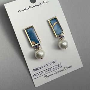 以下が含まれることがあります： 長方形の青と金のアクセント、金色のフレーム、白い真珠のようなドロップが付いたイヤリング。 イヤリングは、日本語のテキストとブランド名「marmor」が記載された白いカードにあります。 カードには、イヤリングがサージカルステンレス鋼であることが示されています。