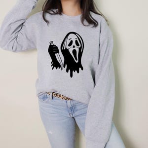 May include: A light gray sweatshirt featuring a black graphic of a ghost-like face holding a knife. The design has a dripping effect. The sweatshirt has long sleeves and a crew neck.