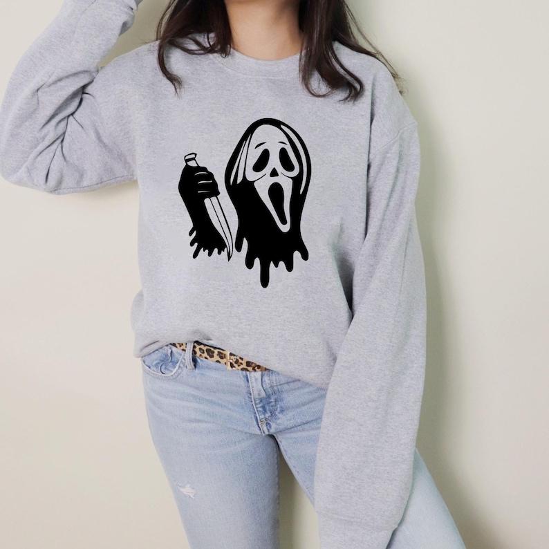 May include: A light gray sweatshirt featuring a black graphic of a ghost-like face holding a knife. The design has a dripping effect. The sweatshirt has long sleeves and a crew neck.