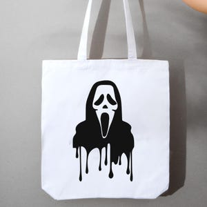 May include: White tote bag featuring a black graphic of the Ghostface mask from the Scream horror film series. The black design has a dripping effect, with the black ink running down the bottom of the bag. The bag has two white handles.