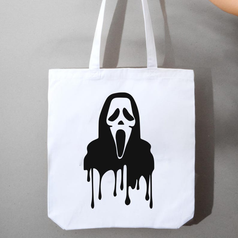 May include: White tote bag featuring a black graphic of the Ghostface mask from the Scream horror film series. The black design has a dripping effect, with the black ink running down the bottom of the bag. The bag has two white handles.