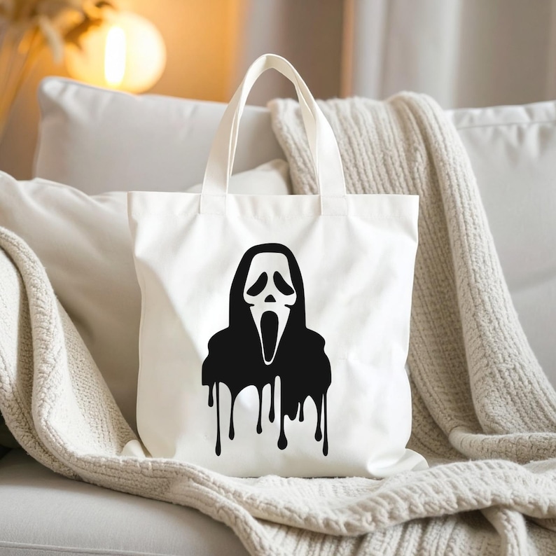 May include: White canvas tote bag featuring a black graphic of the Ghostface mask from the Scream horror film series. The design includes a dripping effect below the mask. The bag has two white handles.