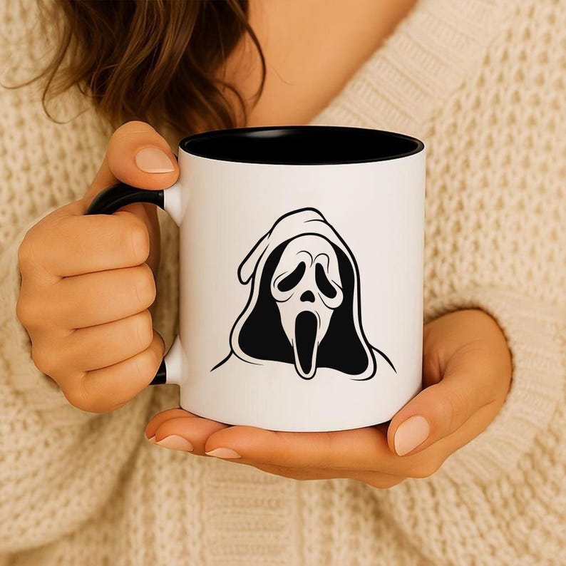 May include: White ceramic mug with a black interior and handle. The mug features a black and white graphic of the Scream mask. The mug is held by a person wearing a cream-colored sweater.