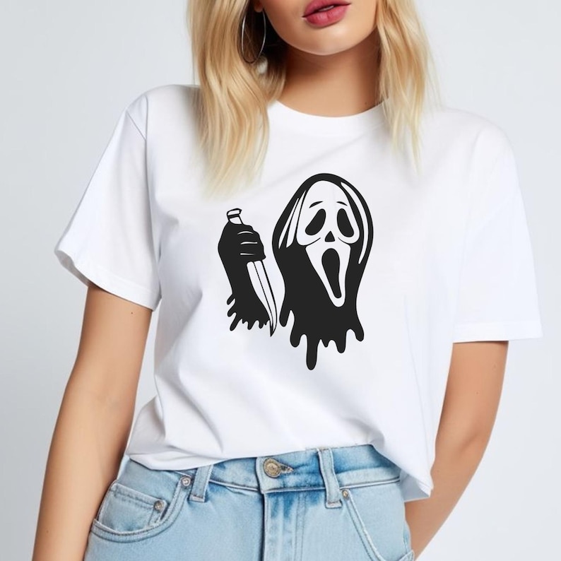 May include: White t-shirt featuring a black graphic of a ghost face holding a knife. The design has a dripping effect. The t-shirt is made of a soft material and has short sleeves. The shirt is a casual wear item.