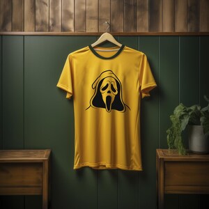 May include: Yellow t-shirt with a black Scream mask graphic. The shirt has a green collar and is hanging on a wooden hanger against a green wall. The design features a hooded figure with an open mouth.