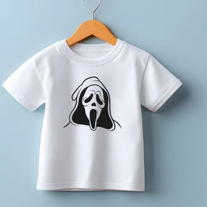 May include: White short-sleeved t-shirt featuring a black and white graphic of a Scream mask. The design is centered on the front of the shirt. The shirt is hanging on a wooden hanger against a light blue background.