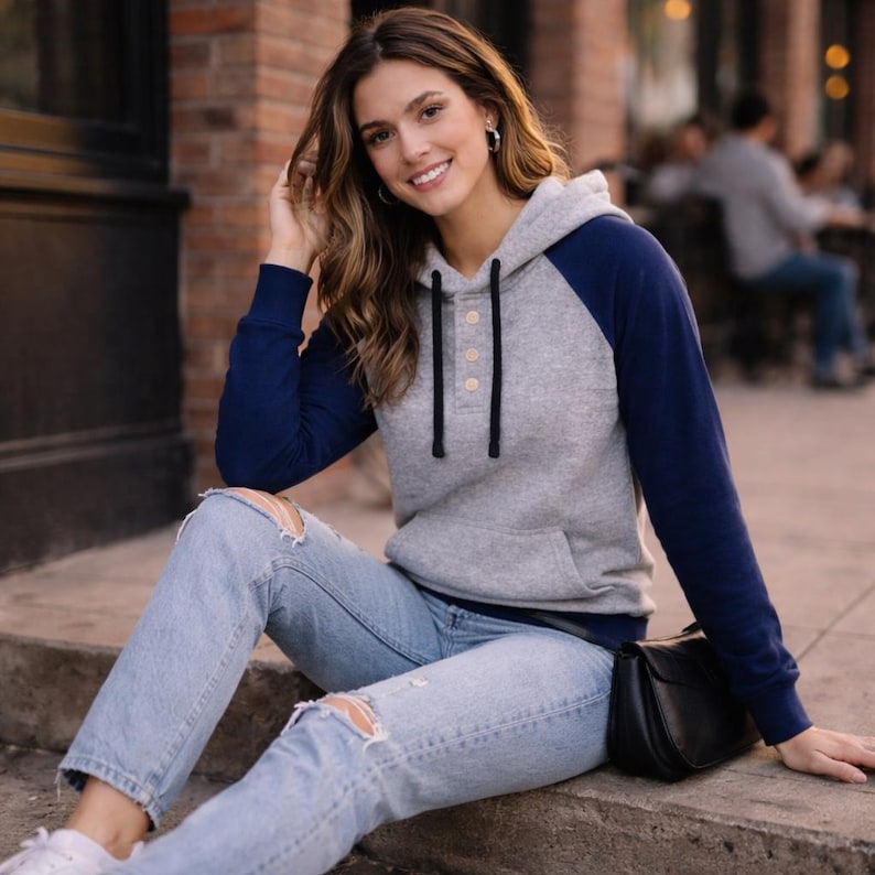 May include: A gray hoodie with navy blue sleeves and a hood. The hoodie has a button-up closure and a front pocket. The person is wearing light blue ripped jeans and white sneakers. A black purse is next to the person.