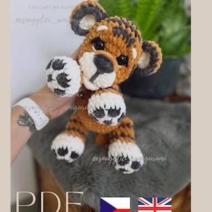 May include: A handmade crochet tiger amigurumi in shades of orange, brown, white, and gray. The tiger has a detailed face, paws, and striped pattern. The image includes the text "PDF" and the shop name "snugglici_amigurumi".