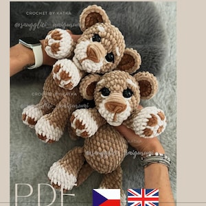 Op de afbeelding: Twee handgemaakte gehaakte leeuw amigurumi speelgoed. De leeuwen zijn bruin en wit met zwarte ogen en bruine neuzen. De tekst "CROCHET BY KATKA" en "@snugglici_amigurumi" zijn zichtbaar. De afbeelding bevat ook de tekst "PDF" en de vlaggen van Tsjechië en het Verenigd Koninkrijk.