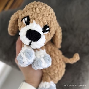 May include: A crocheted puppy amigurumi in shades of tan, white, black, and light blue. The handmade plush toy features floppy ears, a white muzzle, and blue paws. The text "PDF" and "snugglici_amigurumi" are visible.