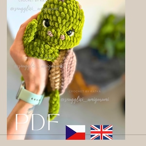 May include: A green and brown crocheted amigurumi dinosaur with black eyes and small pink accents. The text "PDF" is visible, along with the text "snuggllici_amigurumi" and "CROCHET BY KATKA".