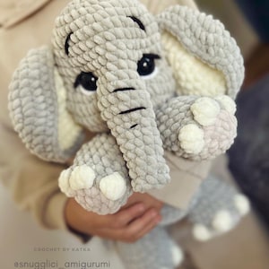 May include: A handmade gray crochet elephant amigurumi with black eyes and a trunk. The elephant has large ears with white accents and white details on its feet. The text "CROCHET BY KATKA" and "@snugglici_amigurumi" are visible.