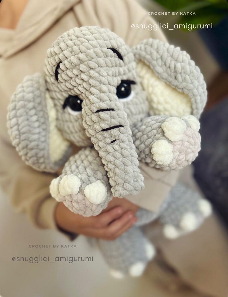 May include: A handmade gray crochet elephant amigurumi with black eyes and a trunk. The elephant has large ears with white accents and white details on its feet. The text "CROCHET BY KATKA" and "@snugglici_amigurumi" are visible.