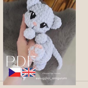 May include: A light gray crocheted cat amigurumi with black eyes and pink accents. The handmade plush toy has a fluffy texture and is shown against a blurred background. The word "PDF" is visible, along with the text "CROCHET BY KATKA" and the Instagram handle @snugglici_amigurumi.