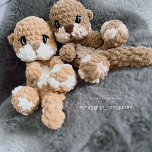 Pu&ograve; includere: Due peluche di lontra all'uncinetto fatti a mano nei toni del beige e del crema, su una superficie grigia e soffice. Le lontre hanno occhi neri e nasi marroni. Il testo "CROCHET BY KATKA" e "@snugglici_amigurumi" sono visibili.