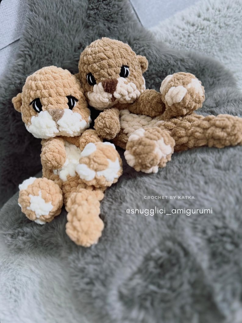 Pu&ograve; includere: Due peluche di lontra all'uncinetto fatti a mano nei toni del beige e del crema, su una superficie grigia e soffice. Le lontre hanno occhi neri e nasi marroni. Il testo "CROCHET BY KATKA" e "@snugglici_amigurumi" sono visibili.