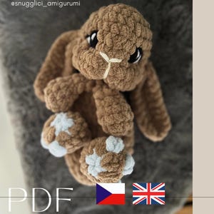 Peut inclure: Un lapin en peluche crocheté marron avec de longues oreilles, des yeux en plastique noir et des accents bleu clair sur les pattes. Le texte CROCHET BY KATKA, @snugglici_amigurumi, PDF et snugglici_amigurumi est visible avec des drapeaux tchèque et britannique.