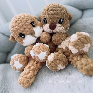 Pu&ograve; includere: Due amigurumi di lontra all'uncinetto fatti a mano. Le lontre sono marroni con accenti bianchi sui loro volti e zampe. I giocattoli sono realizzati con un filato strutturato e hanno occhi neri. Lo sfondo &egrave; un tessuto blu chiaro e morbido. Il testo "CROCHET BY KATKA" e "@snugglici_amigurumi" sono visibili.