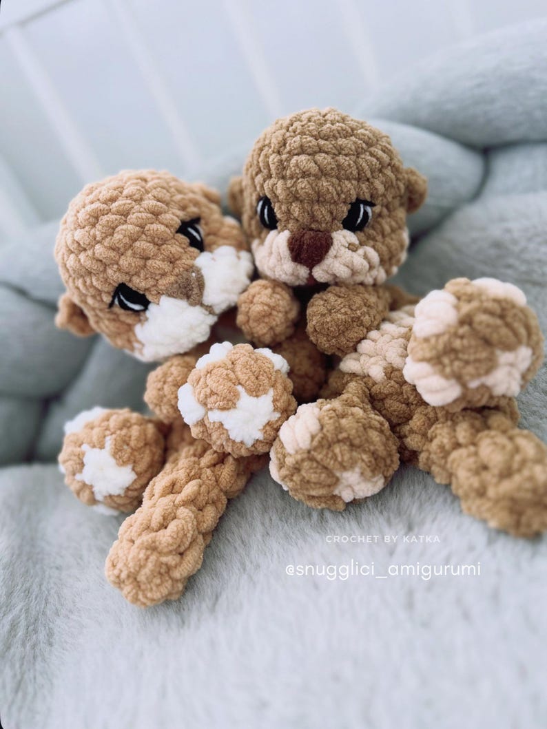 Pu&ograve; includere: Due amigurumi di lontra all'uncinetto fatti a mano. Le lontre sono marroni con accenti bianchi sui loro volti e zampe. I giocattoli sono realizzati con un filato strutturato e hanno occhi neri. Lo sfondo &egrave; un tessuto blu chiaro e morbido. Il testo "CROCHET BY KATKA" e "@snugglici_amigurumi" sono visibili.