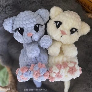 May include: Two handmade crochet cat amigurumi toys. One is light blue, the other is cream-colored. Both have black eyes, pink noses, and pink and blue paw pads. The text "CROCHET BY KATKA" and "@snugglici_amigurumi" are visible.
