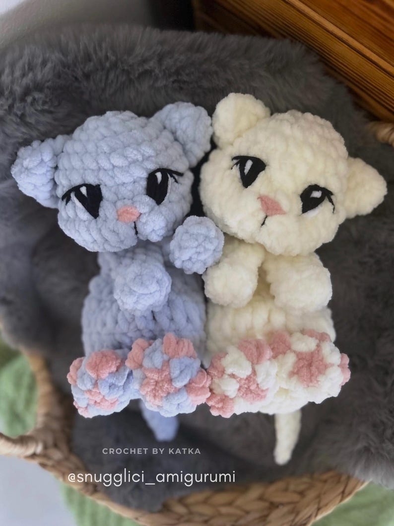 May include: Two handmade crochet cat amigurumi toys. One is light blue, the other is cream-colored. Both have black eyes, pink noses, and pink and blue paw pads. The text "CROCHET BY KATKA" and "@snugglici_amigurumi" are visible.