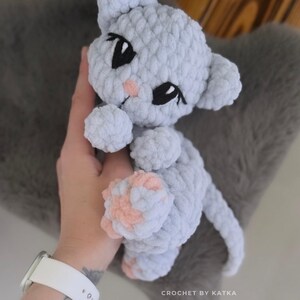 May include: A handmade light gray crochet cat amigurumi with black eyes and pink accents. The cat is held in a hand, and the text "CROCHET BY KATKA" and "@snuggligci_amigurumi" are visible.