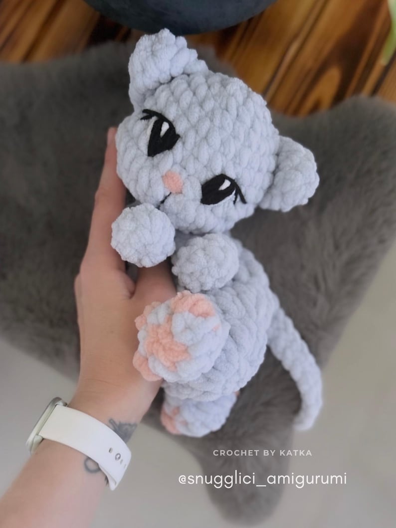 May include: A handmade light gray crochet cat amigurumi with black eyes and pink accents. The cat is held in a hand, and the text "CROCHET BY KATKA" and "@snuggligci_amigurumi" are visible.