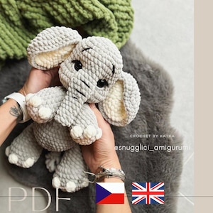 Puede incluir: Un peluche de elefante gris tejido a ganchillo, con orejas grandes y pequeños ojos negros. El elefante se sostiene frente a una manta verde y una superficie gris y peluda. El texto "PDF" y "snugglici_amigurumi" son visibles.