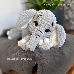 May include: A handmade gray crochet elephant amigurumi with black eyes, white tusks, and pink accents on its feet. The elephant is sitting on a gray faux fur surface. Text on the image reads "CROCHET BY KATKA" and "@snugglici_amigurumi".