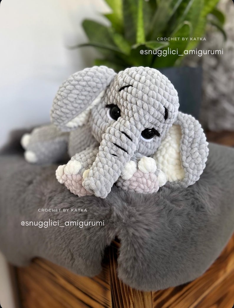 May include: A handmade gray crochet elephant amigurumi with black eyes, white tusks, and pink accents on its feet. The elephant is sitting on a gray faux fur surface. Text on the image reads "CROCHET BY KATKA" and "@snugglici_amigurumi".