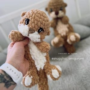 Pu&ograve; includere: Amigurumi di lontra all'uncinetto, marrone e bianco. La lontra ha occhi neri e dettagli bianchi sul petto e sulle zampe. Il testo "CROCHET BY KATKA" e "@snugglici_amigurumi" sono visibili.