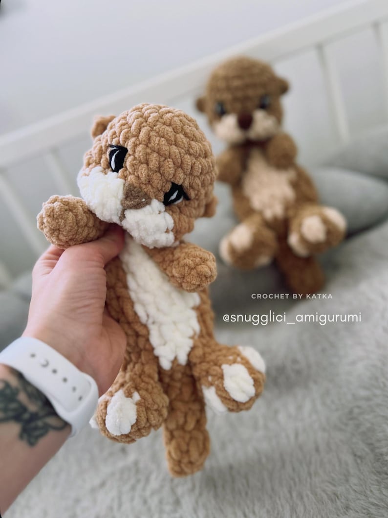 Pu&ograve; includere: Amigurumi di lontra all'uncinetto, marrone e bianco. La lontra ha occhi neri e dettagli bianchi sul petto e sulle zampe. Il testo "CROCHET BY KATKA" e "@snugglici_amigurumi" sono visibili.
