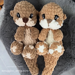 Pu&ograve; includere: Due peluche di lontra fatti a mano all'uncinetto. Le lontre sono marroni e beige con accenti bianchi sulle zampe e sulla pancia. Hanno occhi neri e nasi marroni. I peluche sono su una superficie grigia strutturata. Il testo "CROCHET BY KATKA" e "@snugglici_amigurumi" sono visibili.