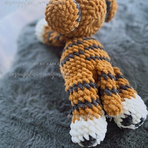 May include: A handmade crochet tiger plush toy in shades of orange, gray, and white. The plush features a striped pattern on its body and head, with white paws and gray accents. The toy is soft and fluffy, perfect for a child's room.