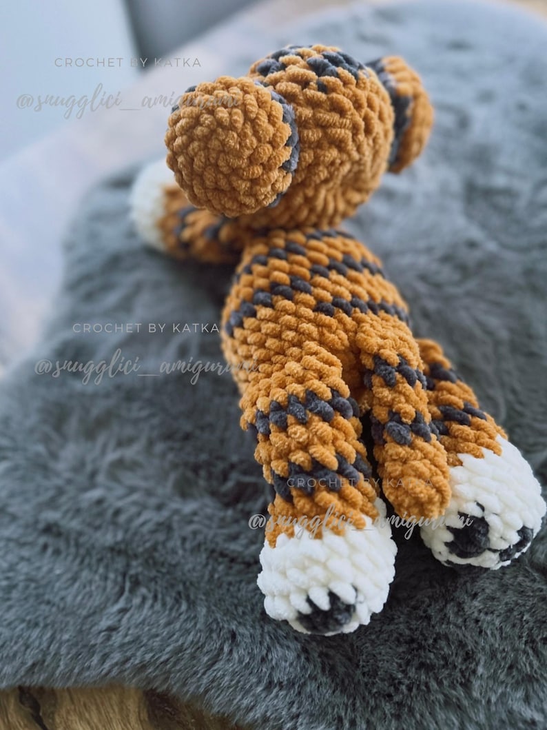 May include: A handmade crochet tiger plush toy in shades of orange, gray, and white. The plush features a striped pattern on its body and head, with white paws and gray accents. The toy is soft and fluffy, perfect for a child's room.