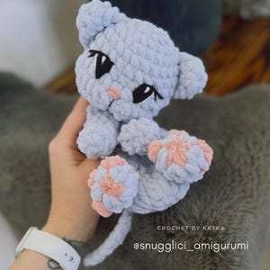 May include: A light blue crocheted cat amigurumi with pink paw pads and black eyes. The handmade plush toy has a small pink nose and is labeled "CROCHET BY KATKA" and "@snugglici_amigurumi".