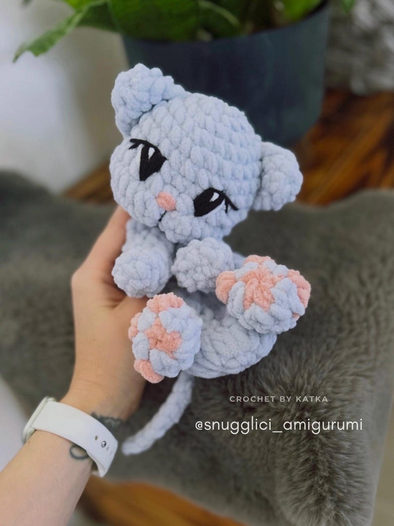 May include: A light blue crocheted cat amigurumi with pink paw pads and black eyes. The handmade plush toy has a small pink nose and is labeled "CROCHET BY KATKA" and "@snugglici_amigurumi".