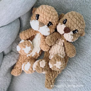 Pu&ograve; includere: Due peluche di lontra all'uncinetto fatti a mano nei toni del marrone e del crema. Le lontre hanno occhi neri e dettagli bianchi sui loro volti e zampe. I peluche sono su una superficie grigia e morbida. Il testo "CROCHET BY KATKA" e "@snugglici_amigurumi" sono visibili.