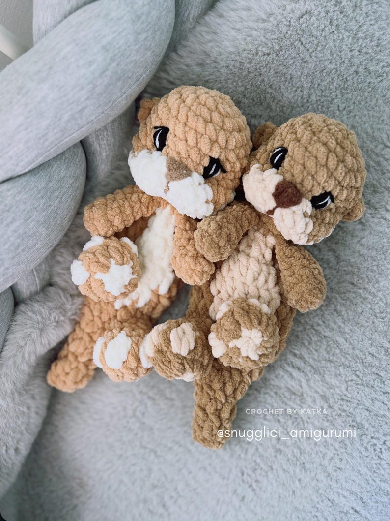 Pu&ograve; includere: Due peluche di lontra all'uncinetto fatti a mano nei toni del marrone e del crema. Le lontre hanno occhi neri e dettagli bianchi sui loro volti e zampe. I peluche sono su una superficie grigia e morbida. Il testo "CROCHET BY KATKA" e "@snugglici_amigurumi" sono visibili.
