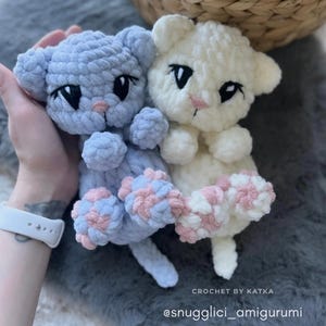 May include: Two handmade crochet cat amigurumi toys. One is light blue, the other is white. Both have black eyes and pink noses. The paws are a mix of pink and blue. The text "CROCHET BY KATKA" and "@snugglici_amigurumi" are visible.
