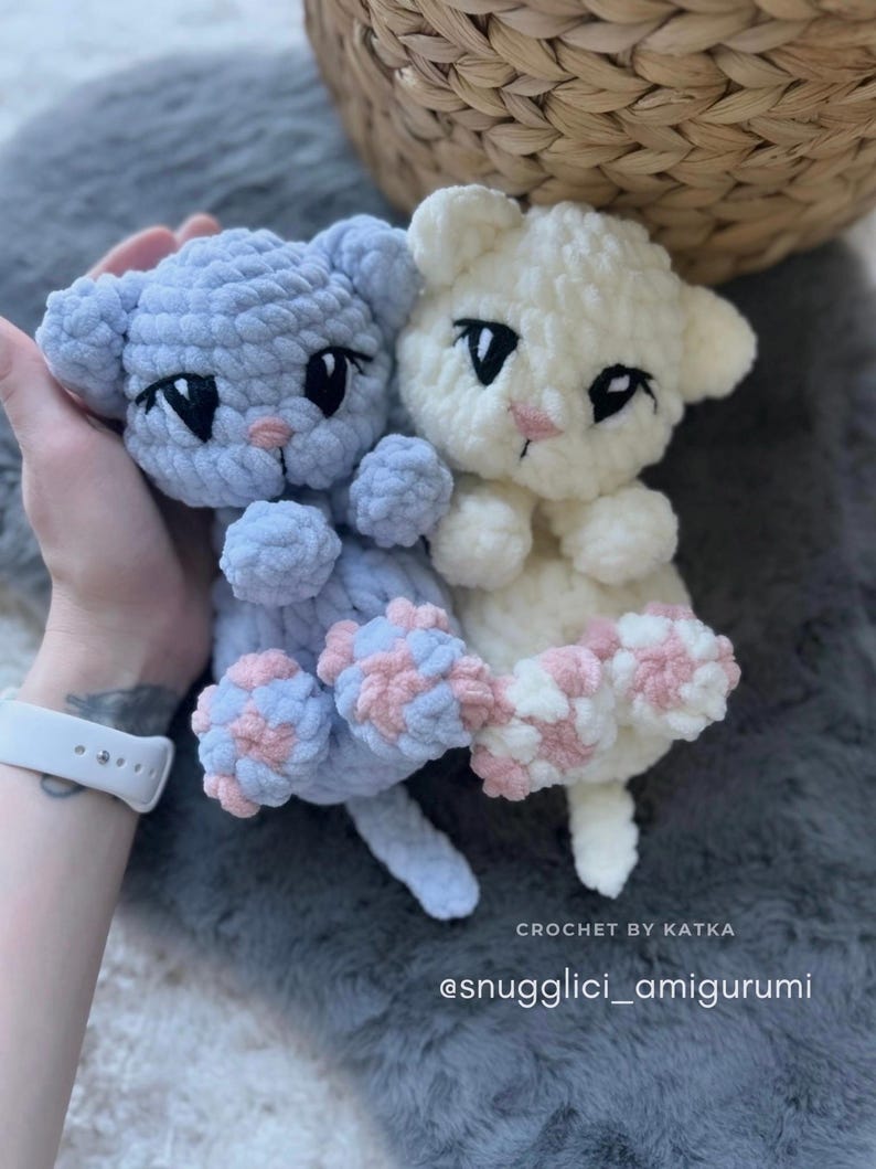 May include: Two handmade crochet cat amigurumi toys. One is light blue, the other is white. Both have black eyes and pink noses. The paws are a mix of pink and blue. The text "CROCHET BY KATKA" and "@snugglici_amigurumi" are visible.