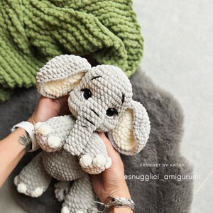 May include: A gray crocheted elephant amigurumi with large ears and black eyes. The elephant is held in hands, with a green blanket in the background. The text "CROCHET BY KATKA" and "@snugglici_amigurumi" are visible.