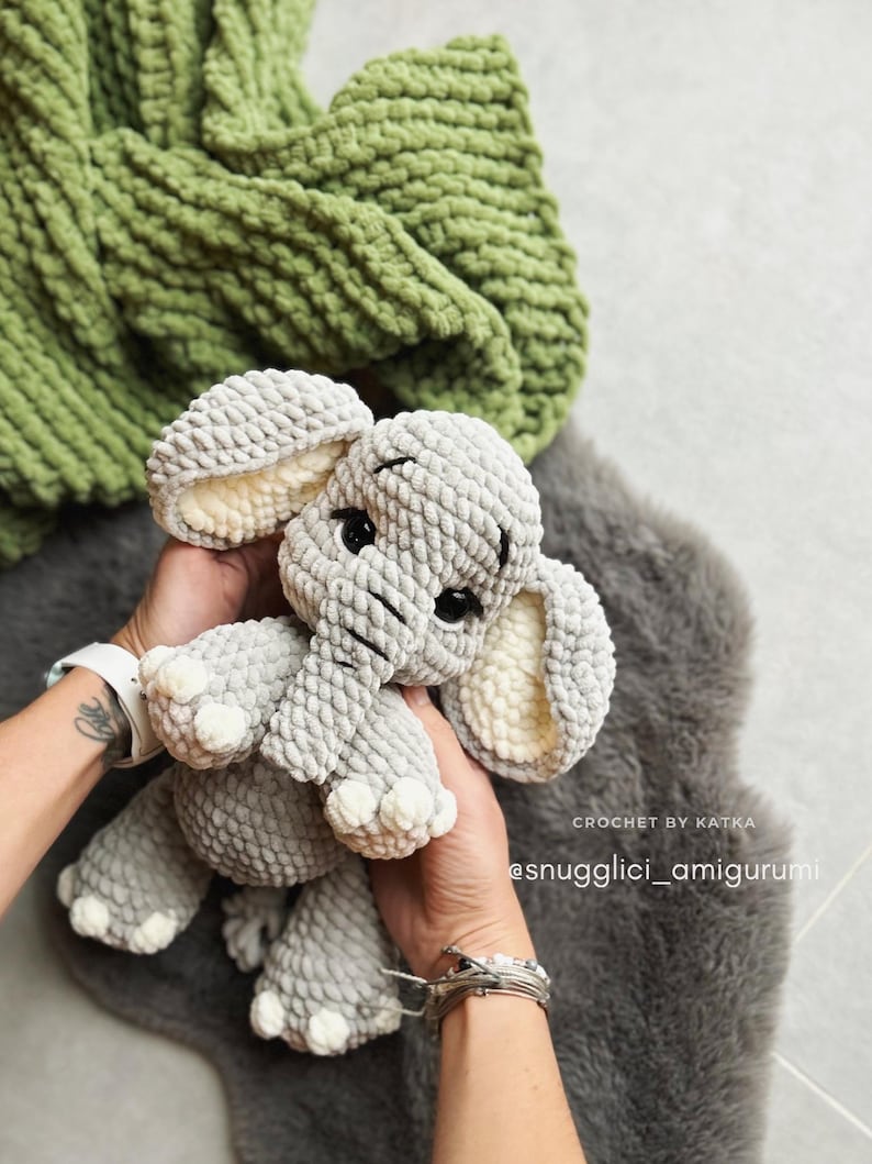 May include: A gray crocheted elephant amigurumi with large ears and black eyes. The elephant is held in hands, with a green blanket in the background. The text "CROCHET BY KATKA" and "@snugglici_amigurumi" are visible.